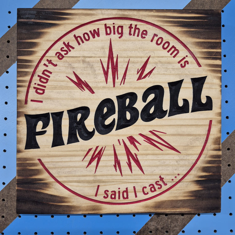 A square 12 by 12 inch solid hardwood carved novelty sign reading "I didn't ask how big the room is, I said I cast FIREBALL" inside a carved fiery explosion. The carving features crisp hand-painted black accents against a blue and brown striped pegboard.