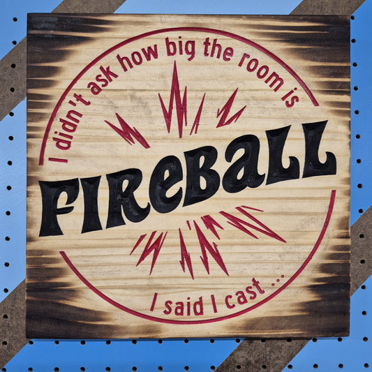 A square 12 by 12 inch solid hardwood carved novelty sign reading "I didn't ask how big the room is, I said I cast FIREBALL" inside a carved fiery explosion. The carving features crisp hand-painted black accents against a blue and brown striped pegboard.