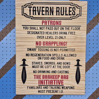 A vertical solid hardwood carved tavern sign featuring funny fantasy rules flanked by two swords. The detailed CNC text features crisp hand-painted black accents against a blue and brown striped pegboard.