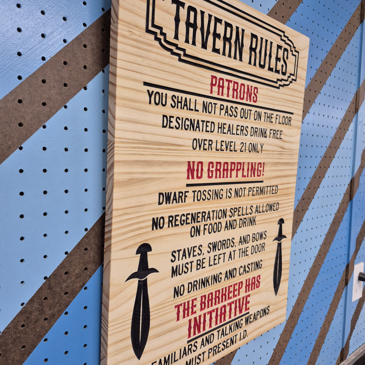 An angled side shot of the custom carved Tavern Rules wooden plaque, highlighting the dimensional depth of the text relief cuts and the rich patterns of the natural hardwood grain against a striped pegboard wall.