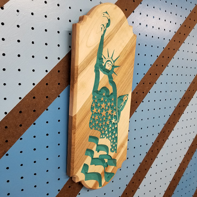 Angled shot of a relief carved hardwood flag sign featuring a detailed Statue of Liberty profile with a star-spangled backdrop painted teal.