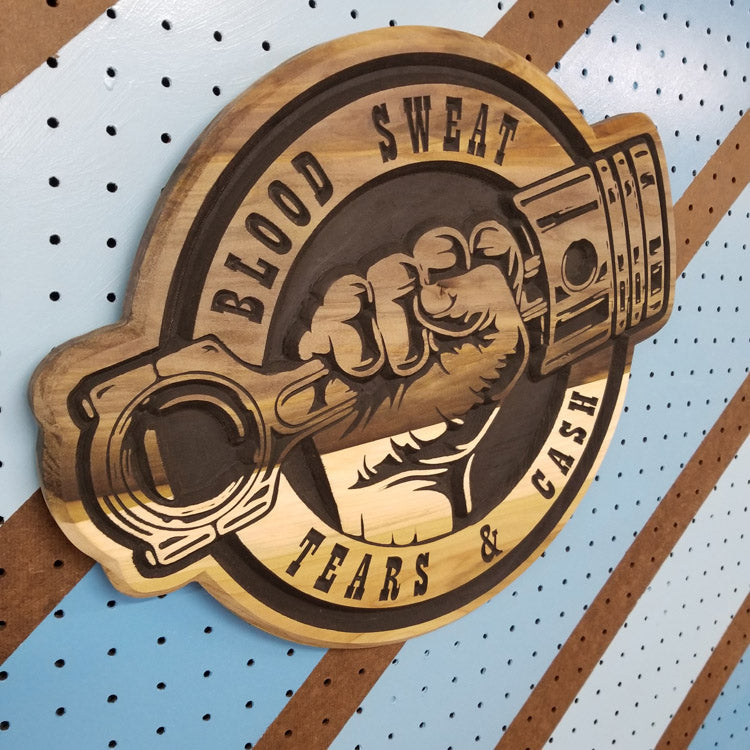 Angled shot of a relief carved hardwood sign with brown painted piston fist design and Blood Sweat Tears & Cash text on pegboard.
