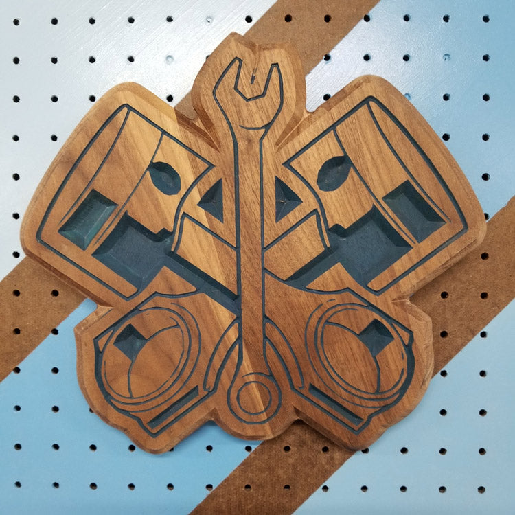 Relief carved hardwood sign with crossed pistons and a wrench design on a striped pegboard.