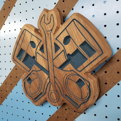 Angled shot of a relief carved hardwood sign with crossed pistons and a wrench design on a striped pegboard.