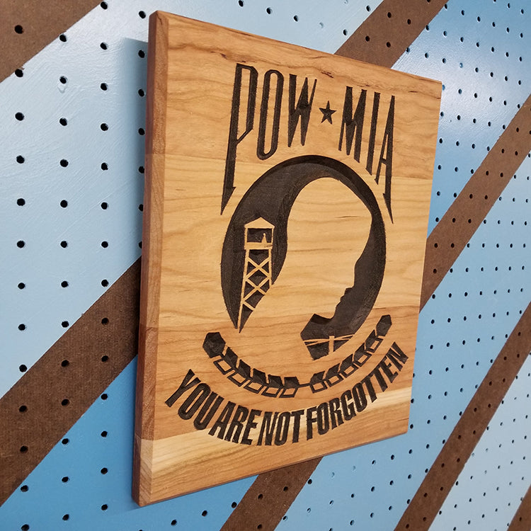Angled shot of a relief carved hardwood POW MIA sign with a silhouette design and You Are Not Forgotten text on a striped pegboard.