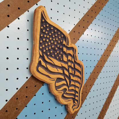 Angled shot of a relief carved hardwood American flag sign with a waving, rippled design on a striped pegboard.
