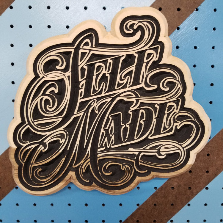 Custom hardwood sign with a detailed, flowing script relief carving of the text Self Made on a striped pegboard.
