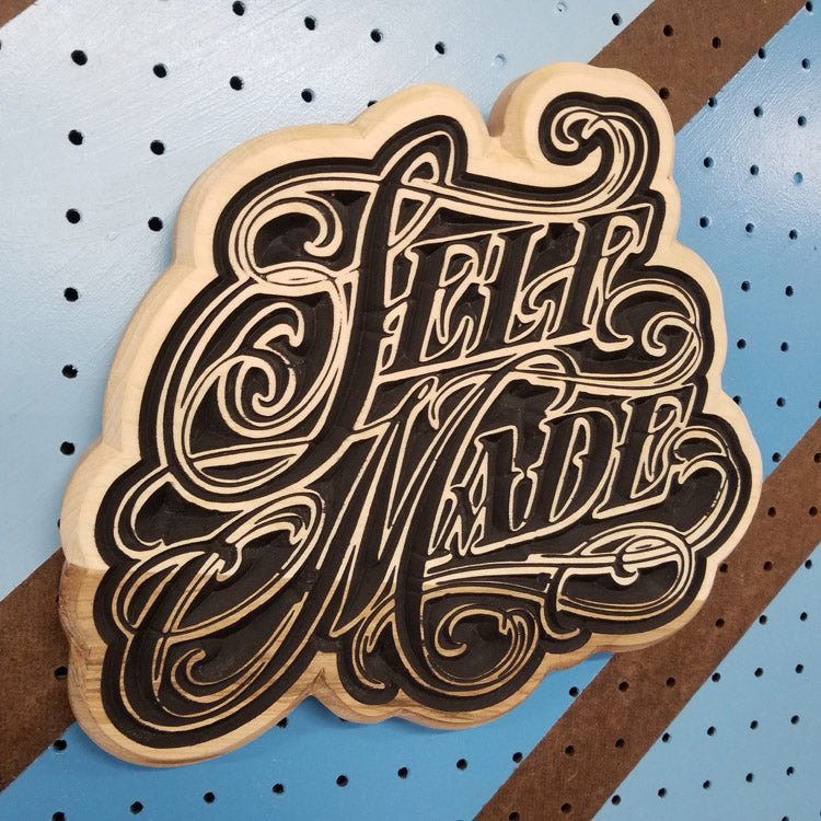 Angled shot of a custom hardwood sign with a detailed, flowing script relief carving of the text Self Made on a striped pegboard.