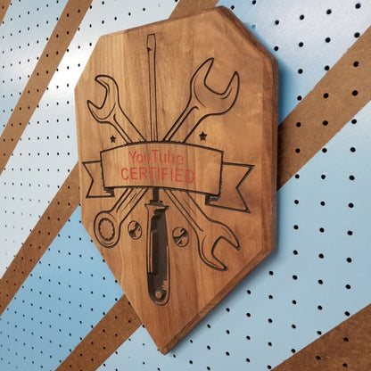 Angled isometric shot of a custom hardwood wrench driver sign with YouTube Certified relief carving on a shop pegboard.